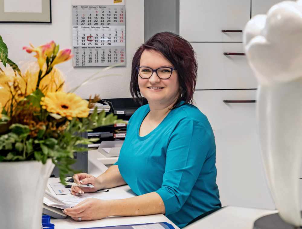 Mitarbeiterin mit türkisfarbener Kleidung am Empfangsschreibtisch, im Hintergrund ein Kalender.