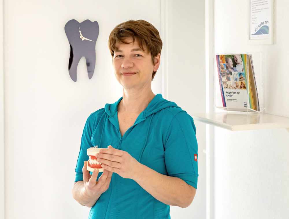 Mitarbeiterin mit türkisfarbener Kleidung hält ein Zahnmodell in der Hand, im Hintergrund eine Zahnform-Uhr und Informationsmaterial.