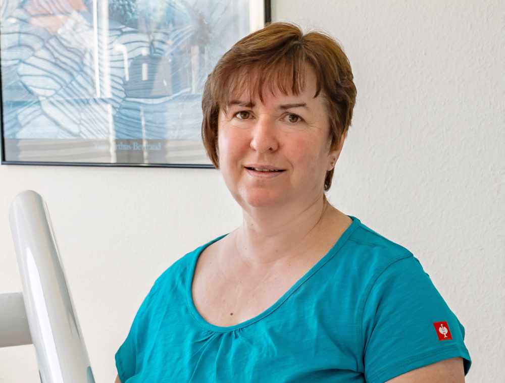 Mitarbeiterin mit türkisfarbener Kleidung in einem Behandlungsraum vor einem Wandbild.