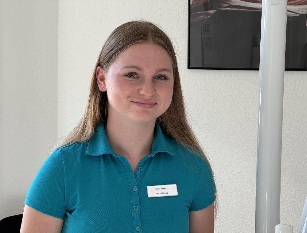 Mitarbeiterin im türkisfarbenen Poloshirt mit Namensschild, lächelnd in einem hellen Innenraum.