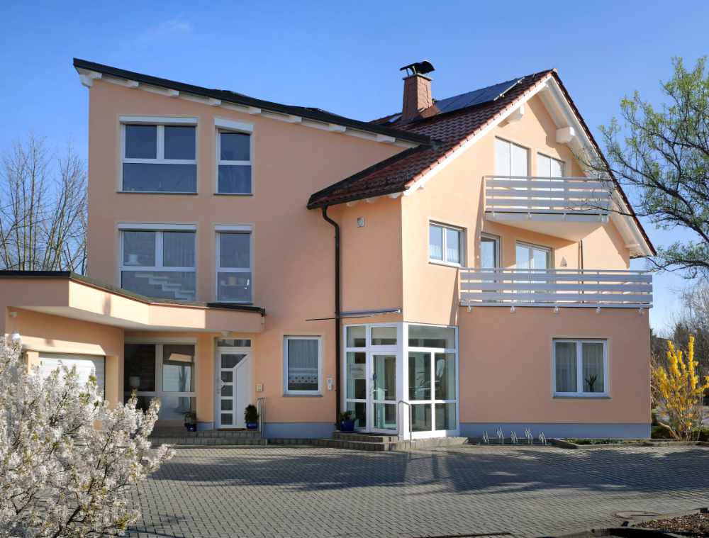 Außenansicht eines hellen Praxisgebäudes mit Eingangsbereich und Balkon, davor ein blühender Baum.