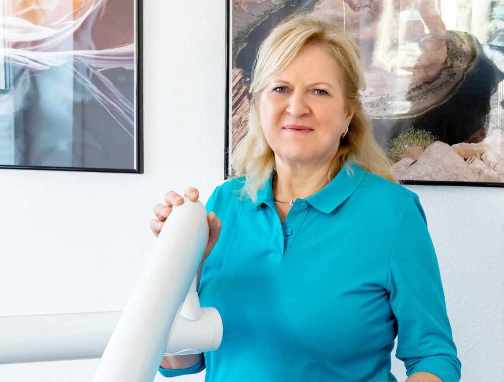 Zahnärztin mittleren Alters mit blondem Haar und türkisfarbener Kleidung steht in einer Zahnarztpraxis neben einem Behandlungsgerät. Im Hintergrund sind zwei gerahmte Bilder an der Wand zu sehen.