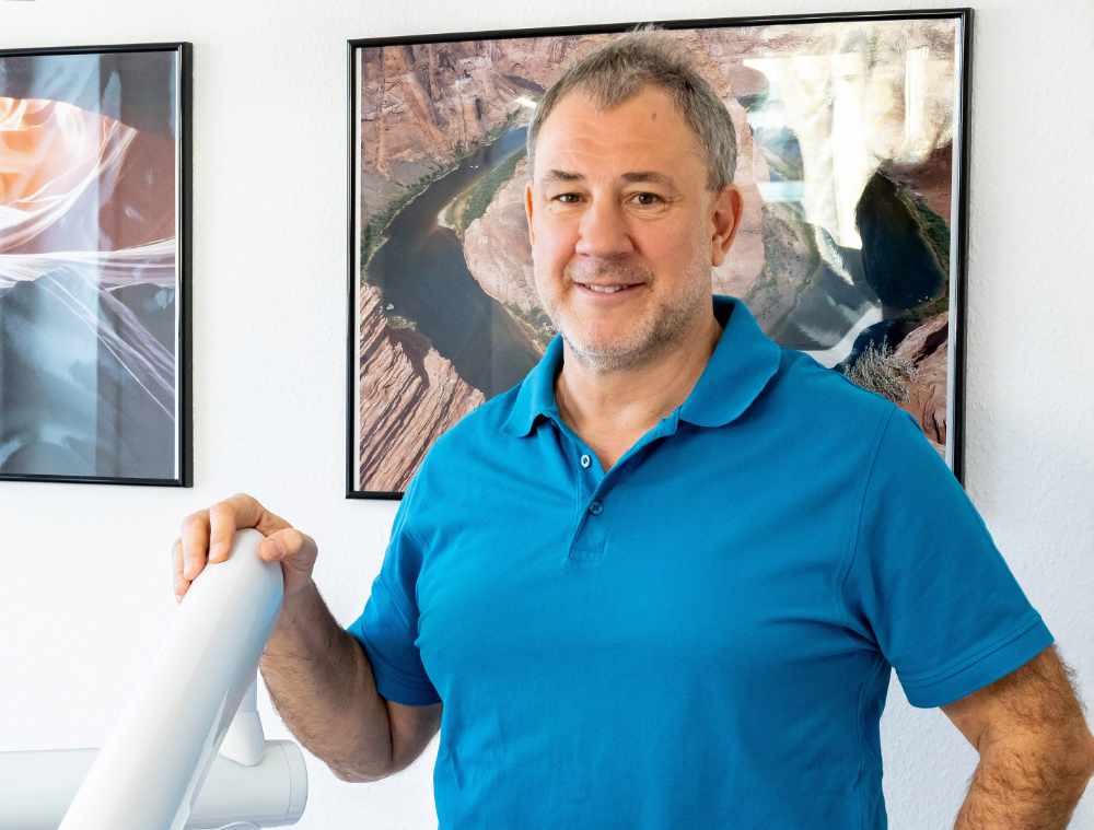 Zahnarzt mit türkisfarbener Kleidung in einem Behandlungsraum vor zwei Wandbildern.