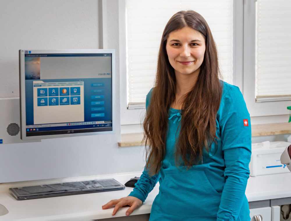 Mitarbeiterin mit türkisfarbener Kleidung vor einem Bildschirm in einem Behandlungsraum.