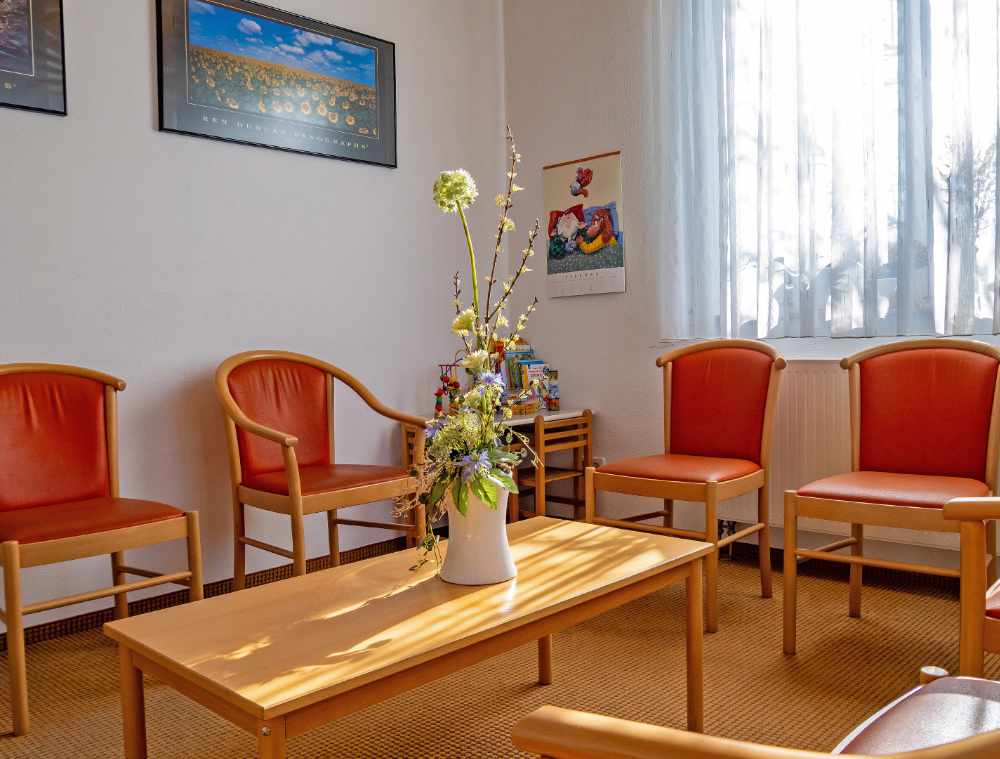 Wartezimmer mit orangefarbenen Stühlen, Holztisch und Blumenvase vor einem Fenster mit Gardine.